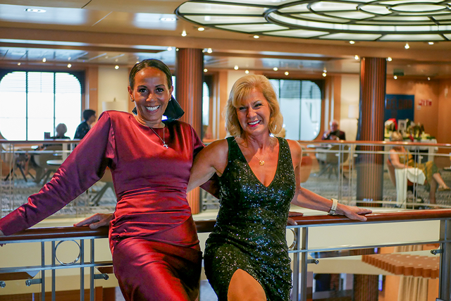 Kerrie Ann and Julie in elegant evening dresses pose together, smiling, on a balcony inside a stylish, well-lit venue.