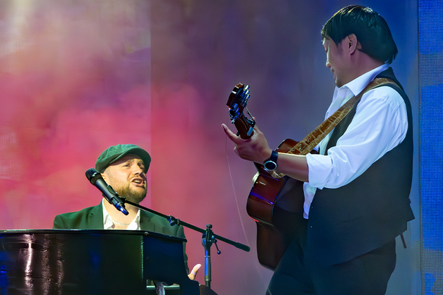 Two musicians perform on stage; one plays the piano and sings, while the other plays an acoustic guitar, set against a colorful backdrop.
