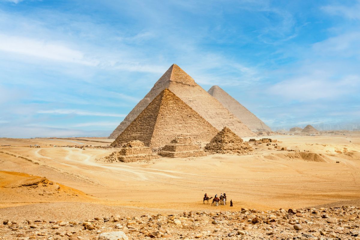 The Pyramids of Giza in Egypt under a blue sky with a group of people and camels in the foreground.