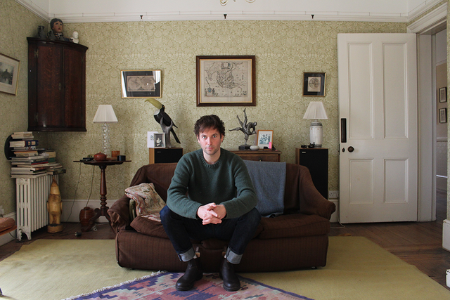 Ben Aitken Photo sitting on a sofa in a vintage-style living room with patterned wallpaper, framed pictures, and decorative items.