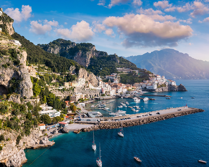 Scenic view of the Amalfi coast with colorful buildings, rugged cliffs, and boats docked in a blue bay under a partly cloudy sky.