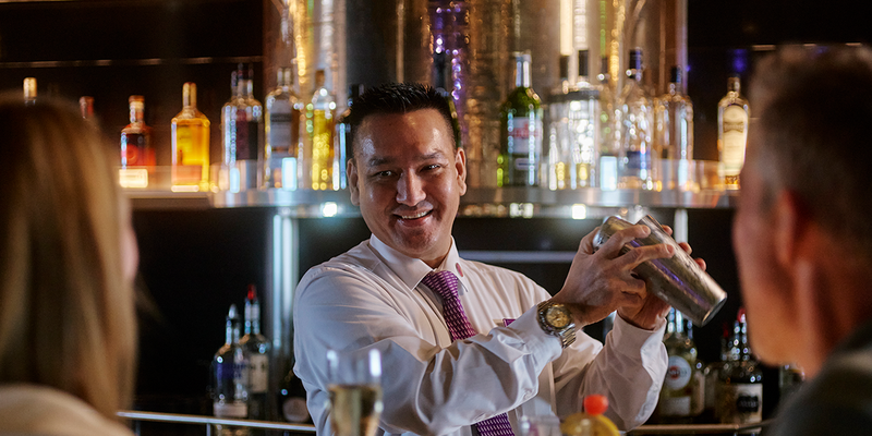 Bartender shaking a cocktail shaker, smiling at guest seated at a bar.