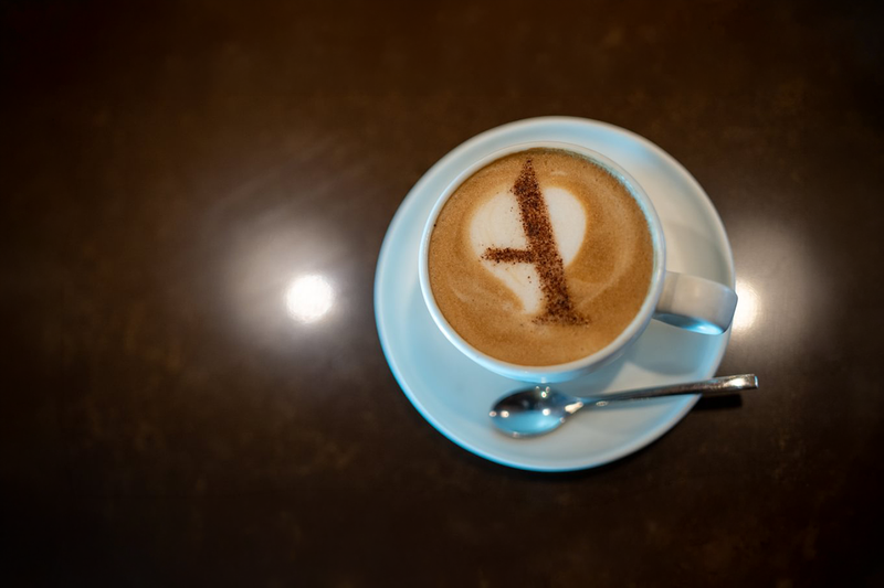 A latte with a letter "A" made of cocoa on the foam, served in a white cup and saucer with a spoon.