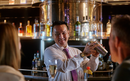 Bartender shaking a cocktail shaker, smiling at guest seated at a bar.