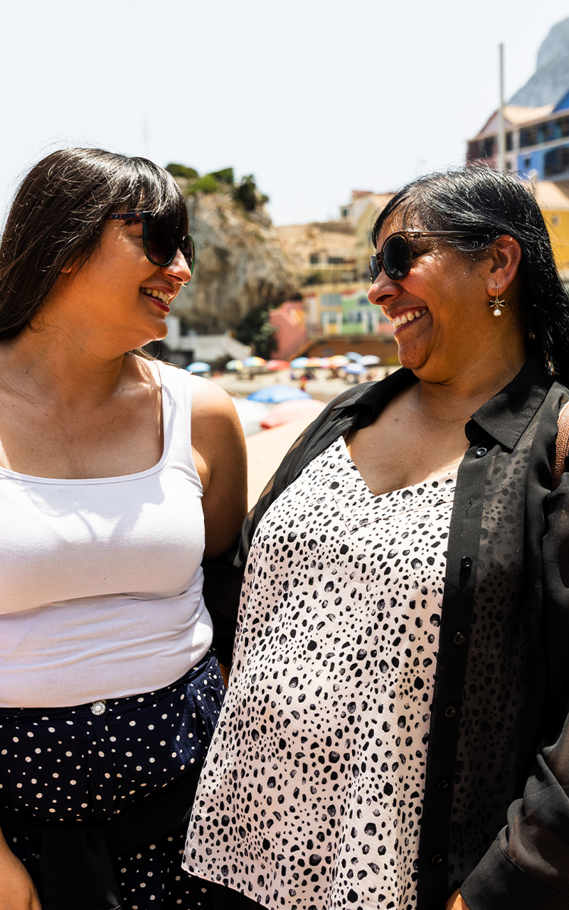 Sarah & Shin smiling at each other outdoors, wearing sunglasses and casual summer attire, with a sunny backdrop of buildings and a beach.