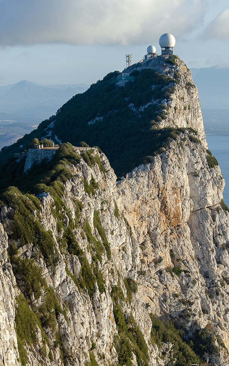 Steep rocky mountain of Gibraltar with radar domes on top, surrounded by lush greenery and a cloudy sky in the background.