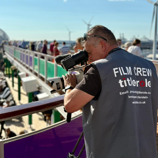 Film crew member with a camera filming on a ship's deck, wearing a vest labeled "FILM CREW titlerole." Passengers and deck chairs are visible.