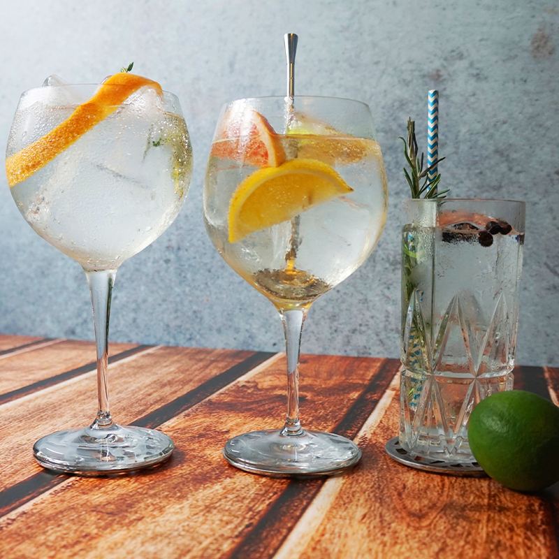 Three cocktails on a wooden surface: two in large glasses with citrus garnishes, and one in a tall glass with herbs and berries, next to a lime.