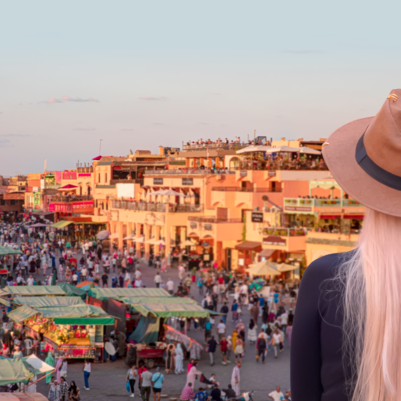 A beautiful blondie young woman in a beige hat watches people in the Jemma el Fnaa or Djemma el Fna famous square in Marrakesh, Morocco