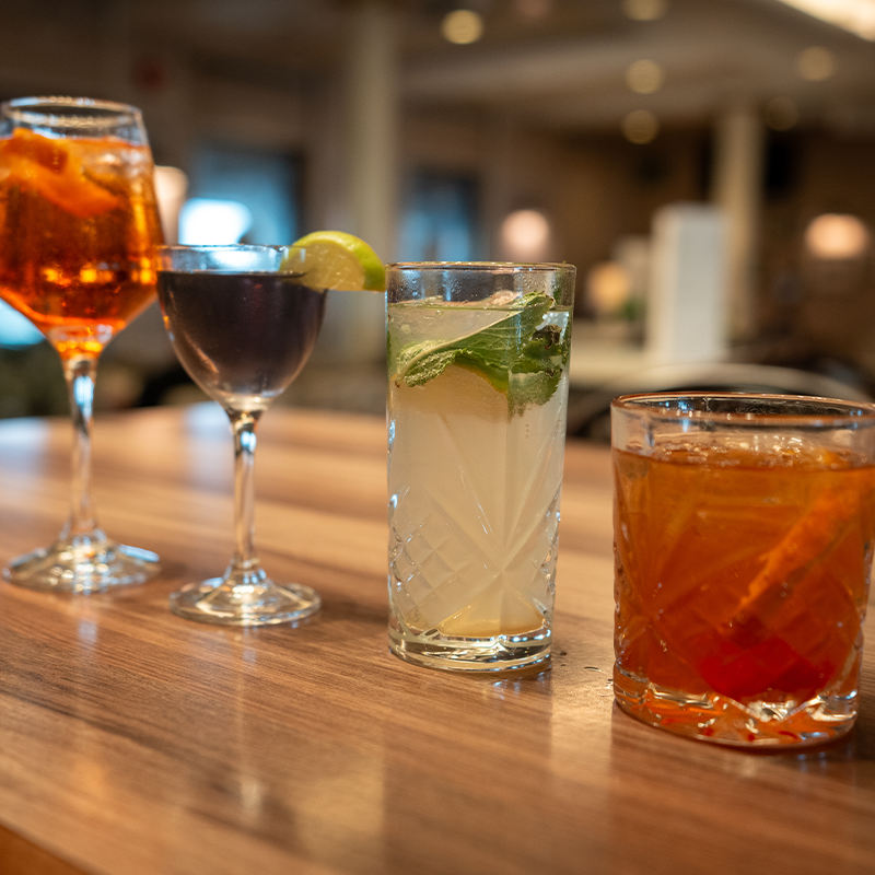 A wooden table displays four cocktails: an orange cocktail, a dark cocktail with lime, a mojito, and a whiskey drink, in a dimly lit room.