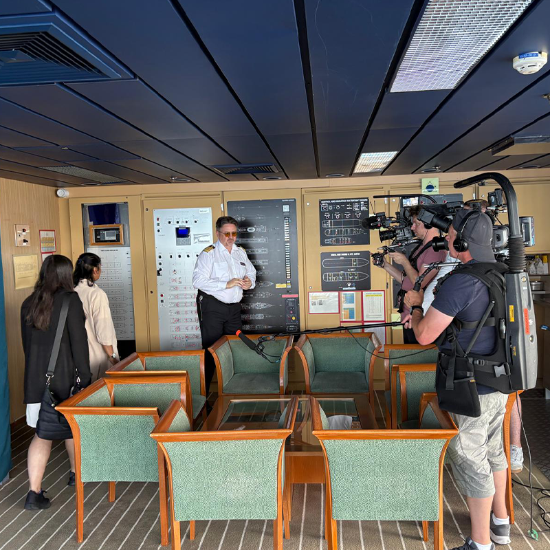 A crew member talks to two people while being filmed by a camera crew in a room with control panels and green chairs.