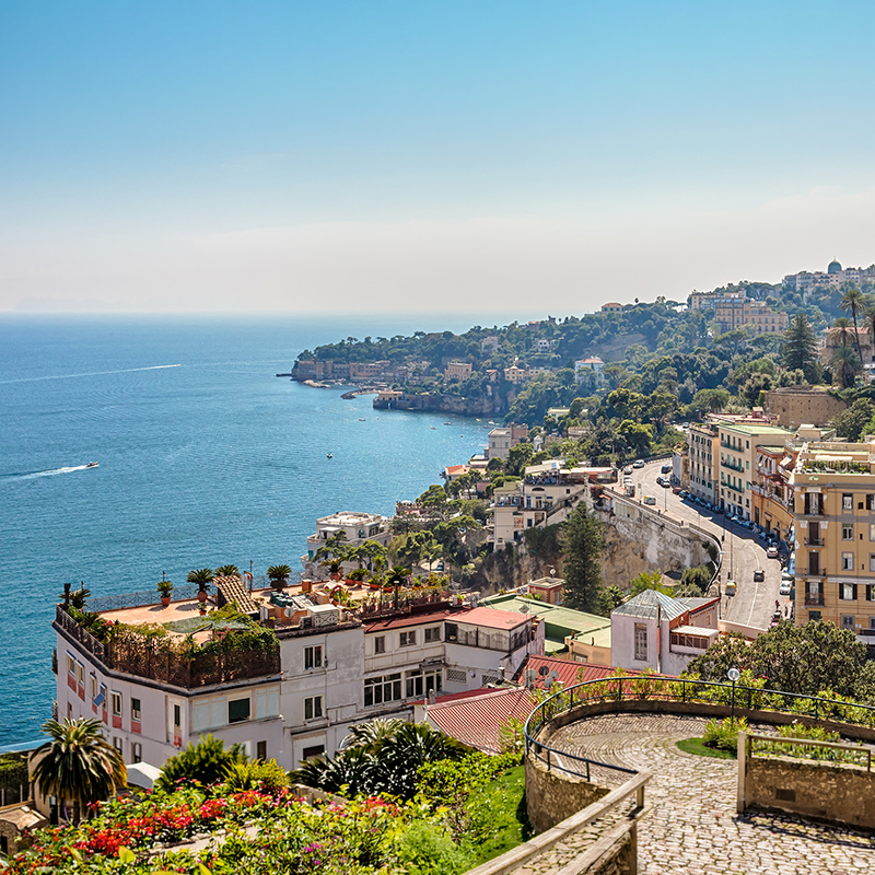 View of a Naples Italy with lush greenery, colorful buildings, and a winding road along a blue sea under a clear sky.